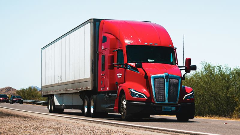 Red semi truck pulling a white dry van trailer on a desert highway under a clear blue sky